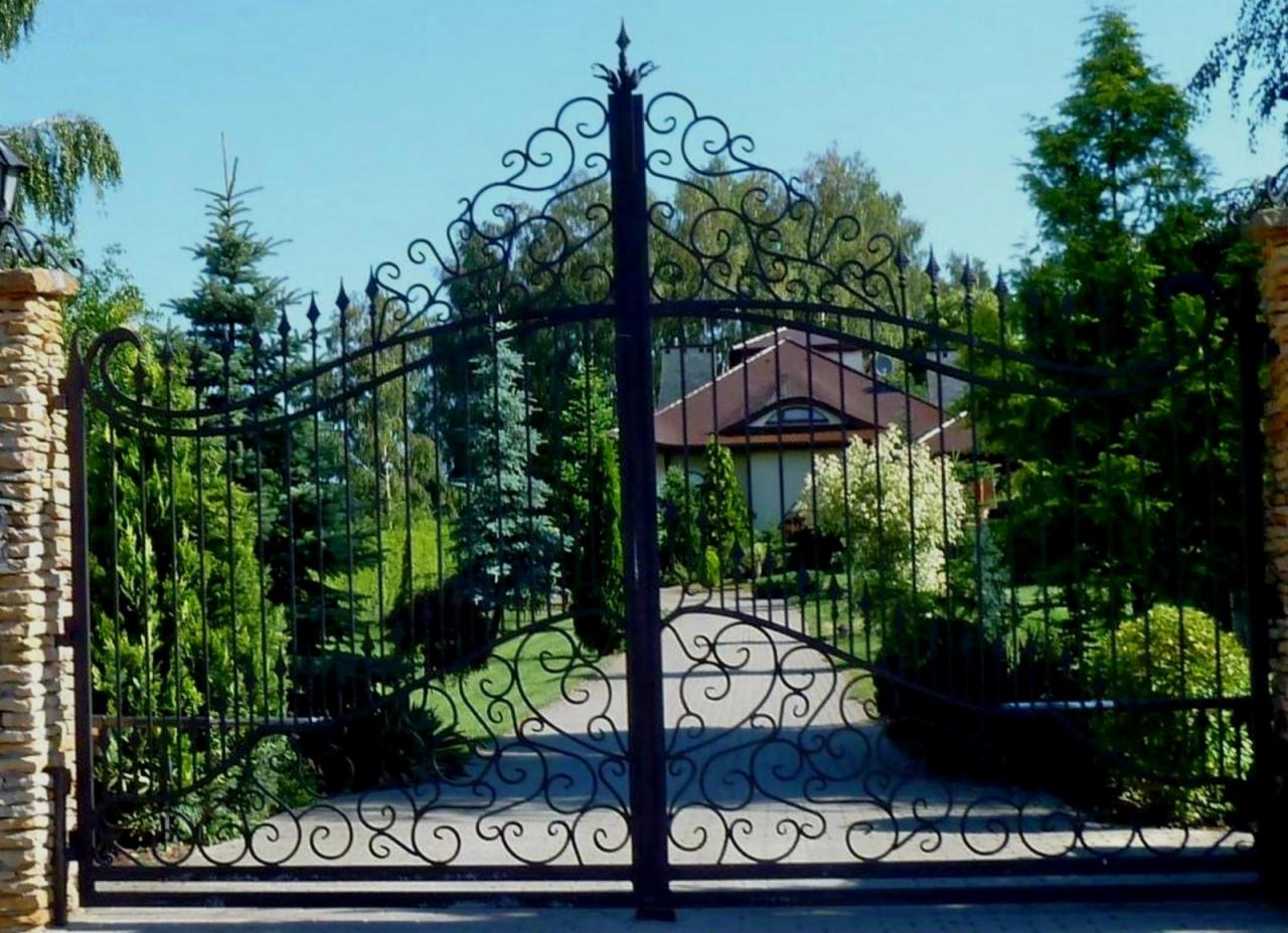 Kuta, ozdobna brama wjazdowa z kamiennymi słupkami, prowadząca do domu otoczonego zielenią. W tle widoczny budynek z czerwonym dachem i zadbany ogród.