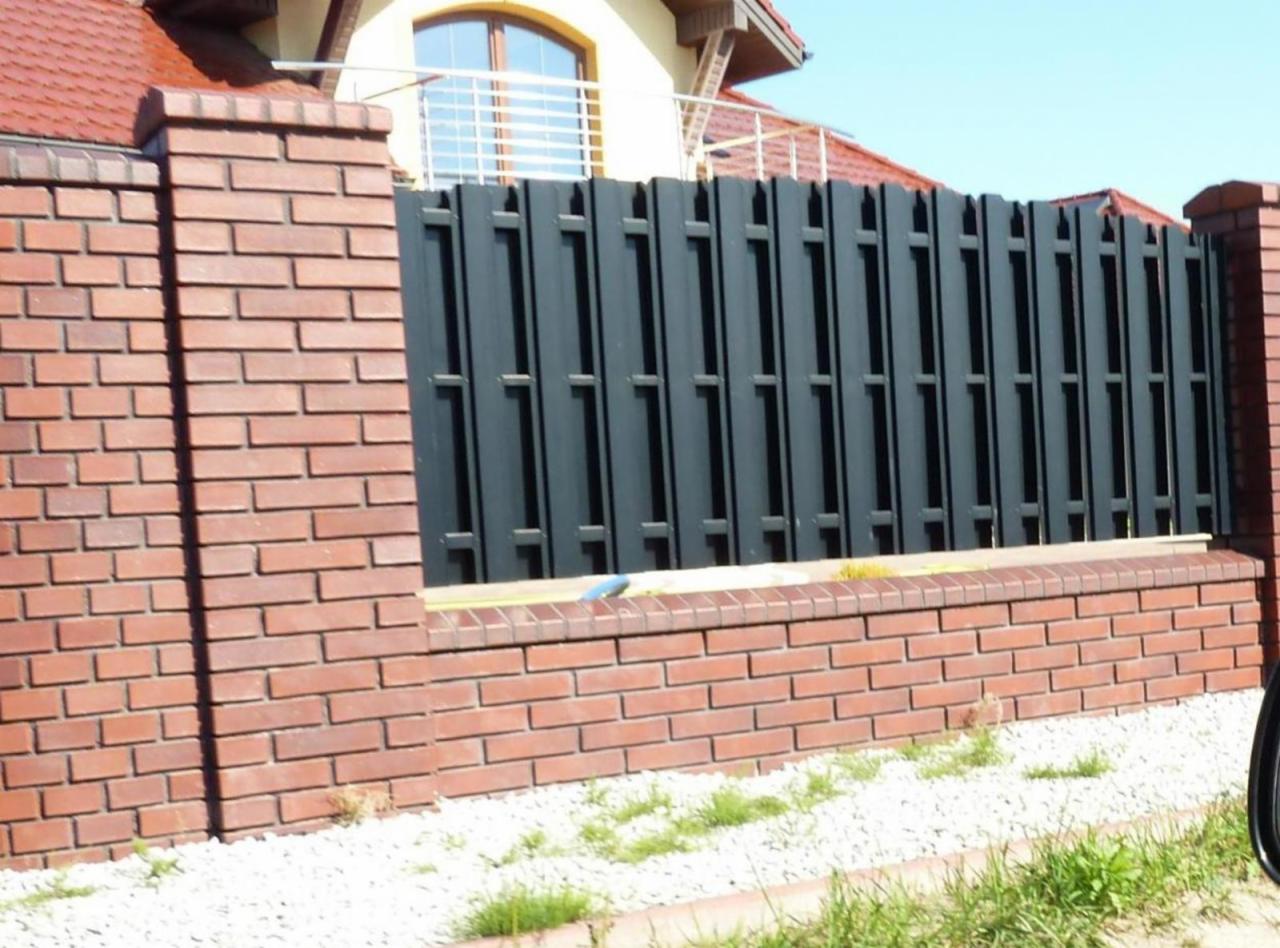 Ciemnoszare, nowoczesne ogrodzenie panelowe z ceglaną podmurówką. Widok z zewnątrz posesji, fragment domu w tle. Elementy zieleni i drobny żwir u podstawy.
