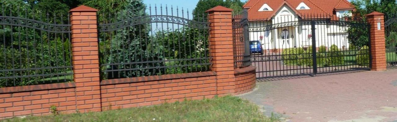 Kuta, czarna brama wjazdowa z ceglanymi słupkami i podmurówką. Brama z ozdobnymi elementami, na tle domu z czerwoną dachówką i zieleni.