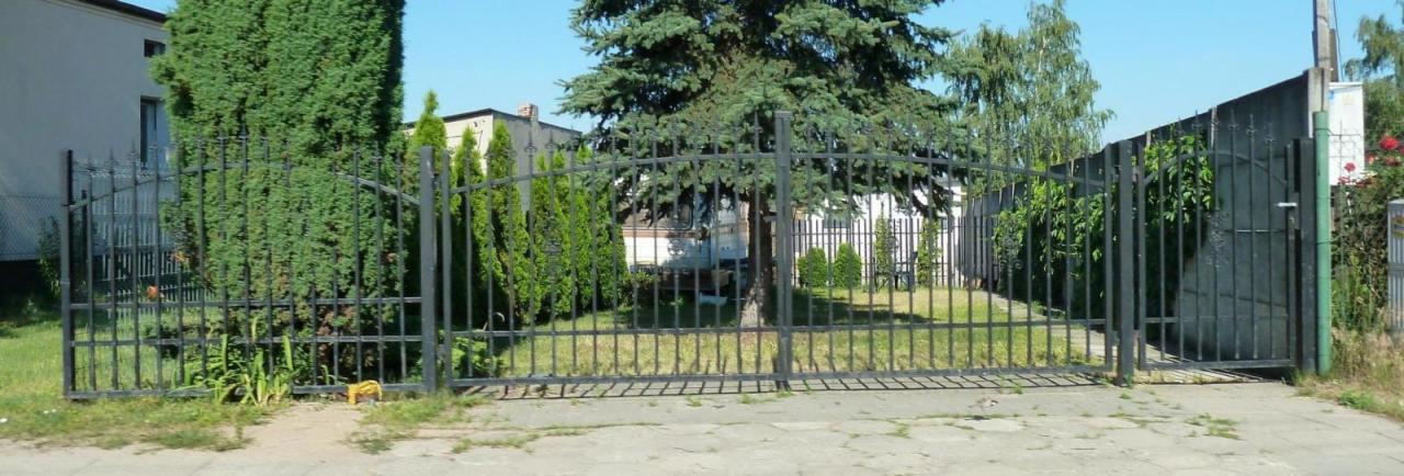 Czarna, metalowa brama wjazdowa z łukowym zwieńczeniem, otoczona zielenią, prowadząca na posesję z domem w tle.