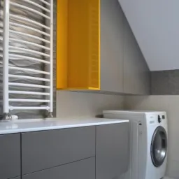 Szare szafki z białym blatem i żółtą otwartą szafką wiszącą w łazience w Łodzi, obok biała pralka.
