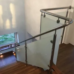 Klatka schodowa z drewnianymi stopniami i balustradą ze stali nierdzewnej z wypełnieniem z matowego szkła, widok z góry na dół, okno z narzędziami na parapecie w tle.