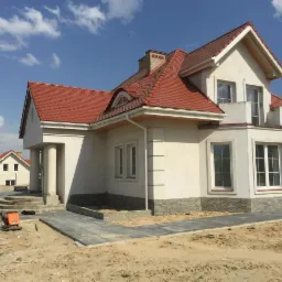 Nowoczesny dom jednorodzinny z jasną elewacją, czerwoną dachówką i kamienną podmurówką, widok z poziomu gruntu na tle błękitnego nieba z chmurami, na pierwszym planie ułożona kostka brukowa i piasek.