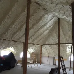 Poddasze po ociepleniu pianką poliuretanową, widoczne drewniane elementy konstrukcyjne, okna dachowe, drabina i materiały budowlane na podłodze.