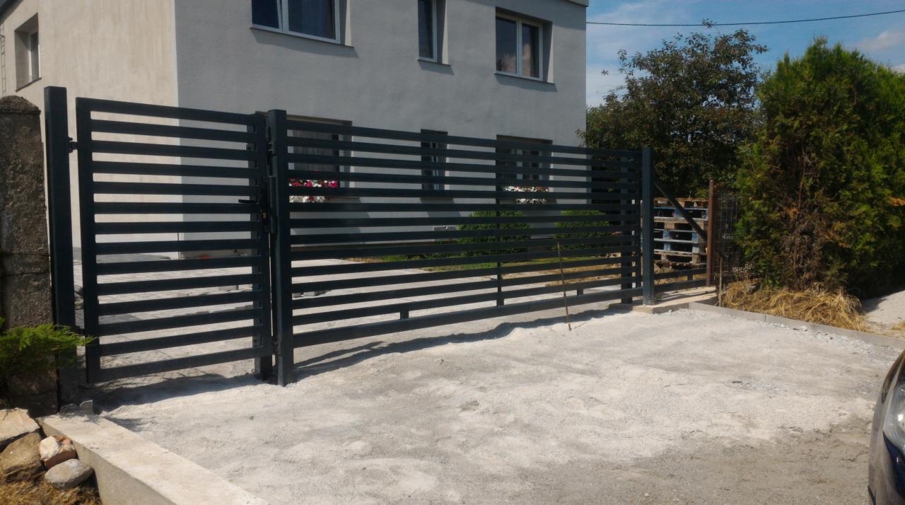 Nowoczesna, pozioma brama panelowa w antracytowym kolorze, z widocznym słupkiem betonowym i fragmentem elewacji budynku w tle. Brama otwarta, wjazd nieutwardzony.