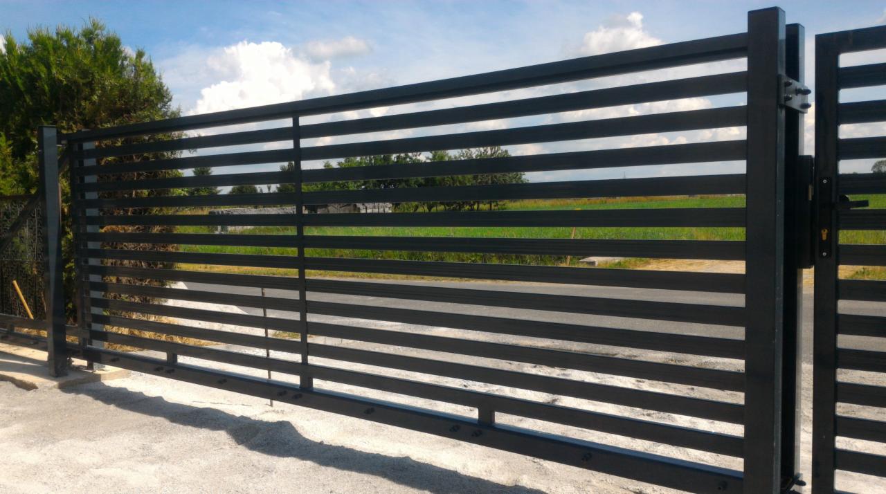 Nowoczesna brama posesyjna z poziomych, metalowych profili w kolorze grafitowym, widok z zewnątrz, z zielonym tłem w oddali i fragmentem ogrodzenia po lewej.