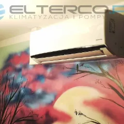 Biały klimatyzator marki Innova zamontowany na ścianie z ręcznie malowanym muralem przedstawiającym drzewa i zachodzące słońce, widoczne logo firmy ELTERCO.PL na górze.