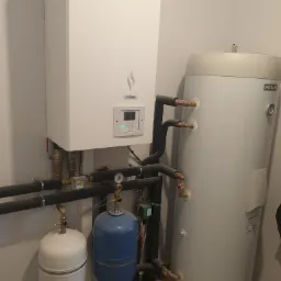 Biała jednostka Yutaki i zbiornik Hitachi z rurami, manometrami i zaworami, zainstalowane przy jasnej ścianie.