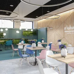 Wnętrze kawiarni z drewnianą ścianą z napisem #kulturalna COFFEE & LUNCH, stoły z kolorowymi krzesłami, rośliny w donicach i okrągłe lampy sufitowe.