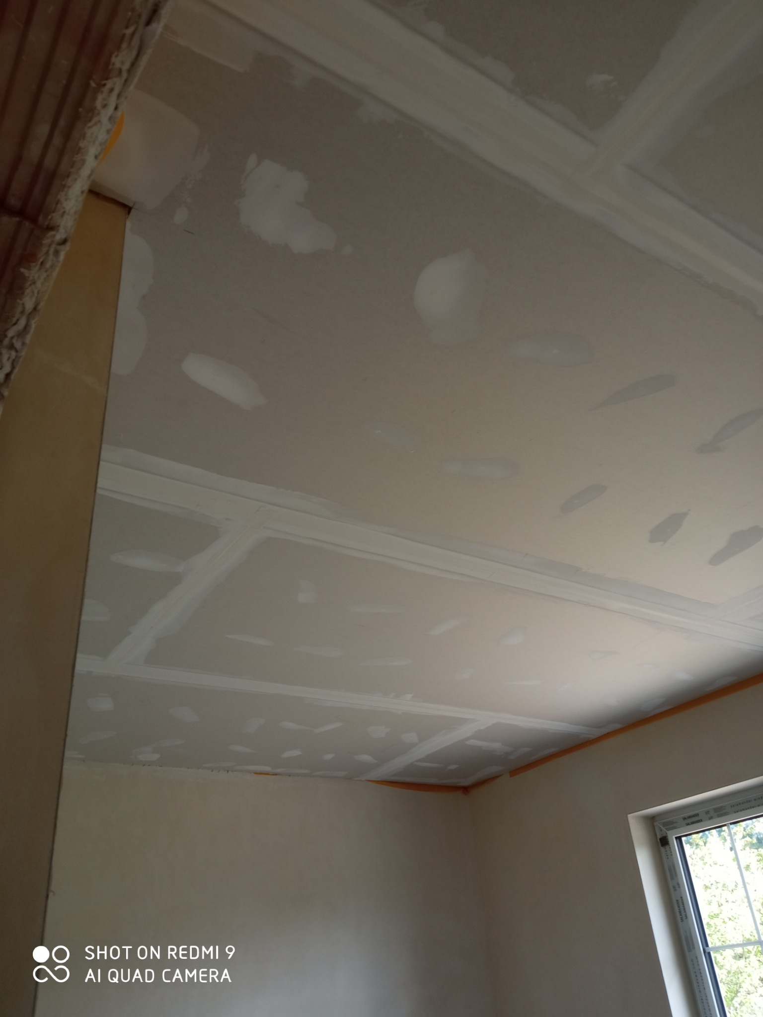 Sufit z płyt kartonowo-gipsowych w trakcie wykańczania, widoczne łączenia wzmocnione siatką i częściowo pokryte szpachlą, narożnik z taśmą ochronną, surowy stan przed malowaniem.