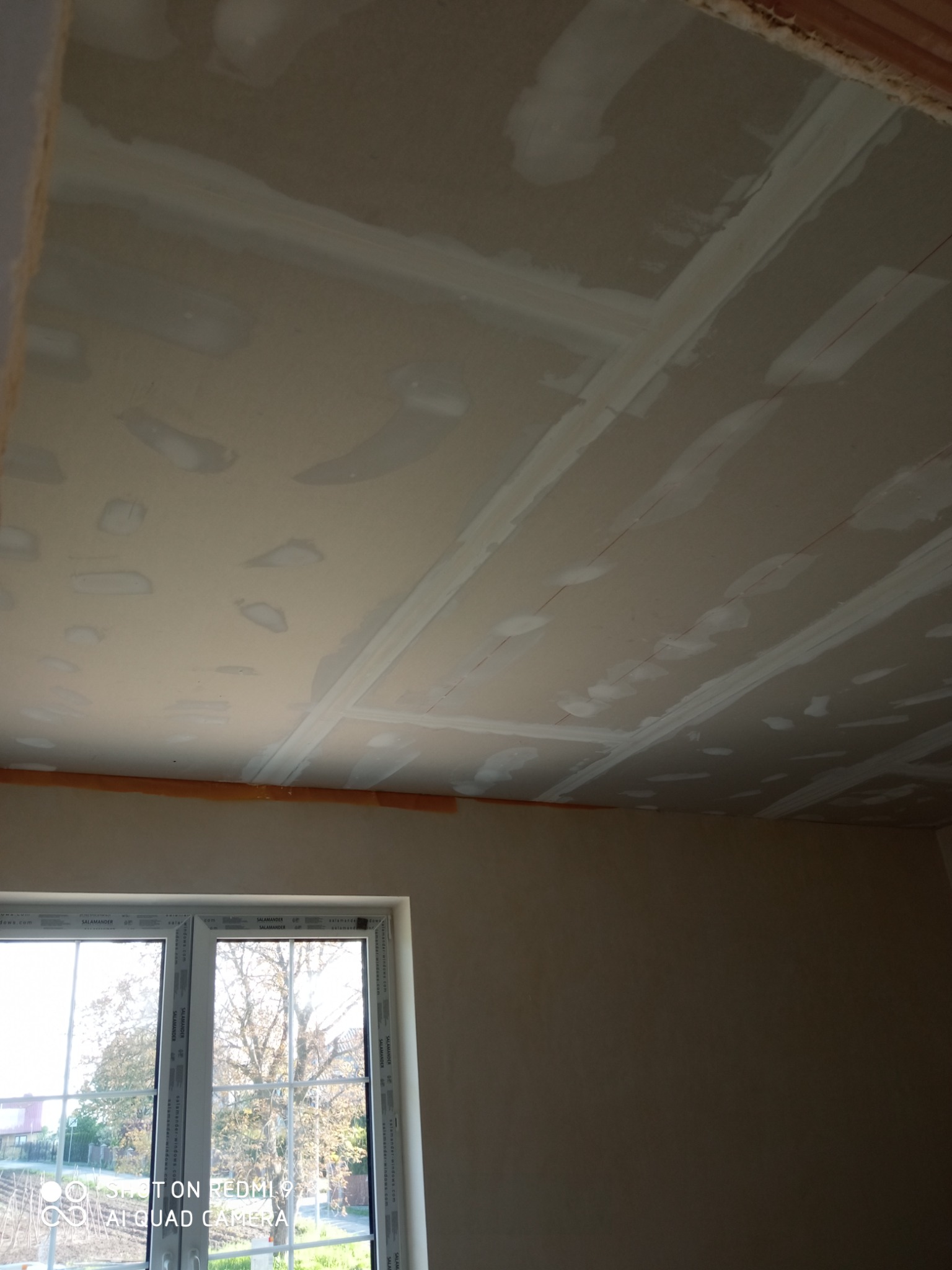 Nierówna powierzchnia sufitu z płyt gipsowo-kartonowych, w trakcie spoinowania łączeń, widoczne niedoskonałości w nałożeniu masy szpachlowej, okno z widokiem na drzewo na zewnątrz.