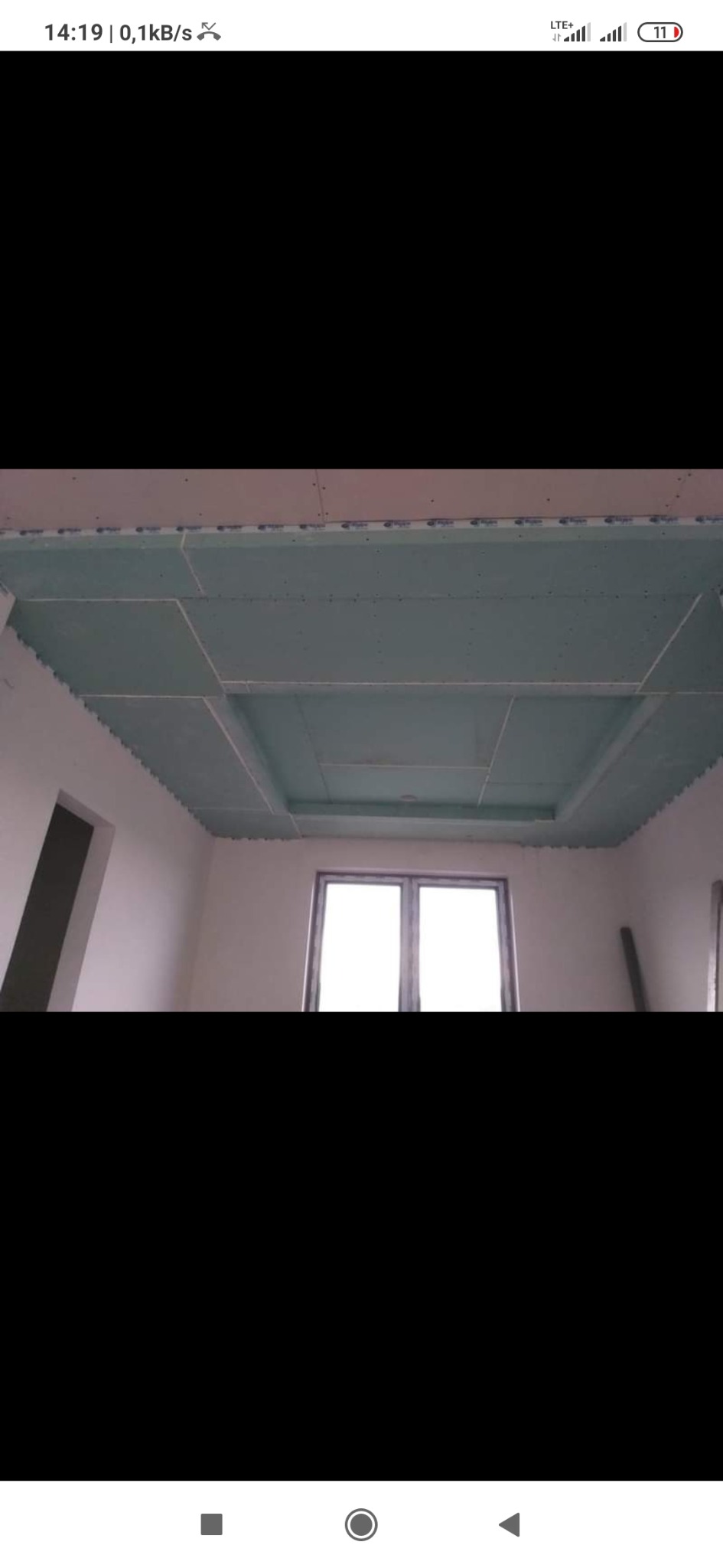 Niedokończony sufit podwieszany z płyt kartonowo-gipsowych z widoczną konstrukcją nośną i niezamalowanymi łączeniami, w pomieszczeniu z oknem.