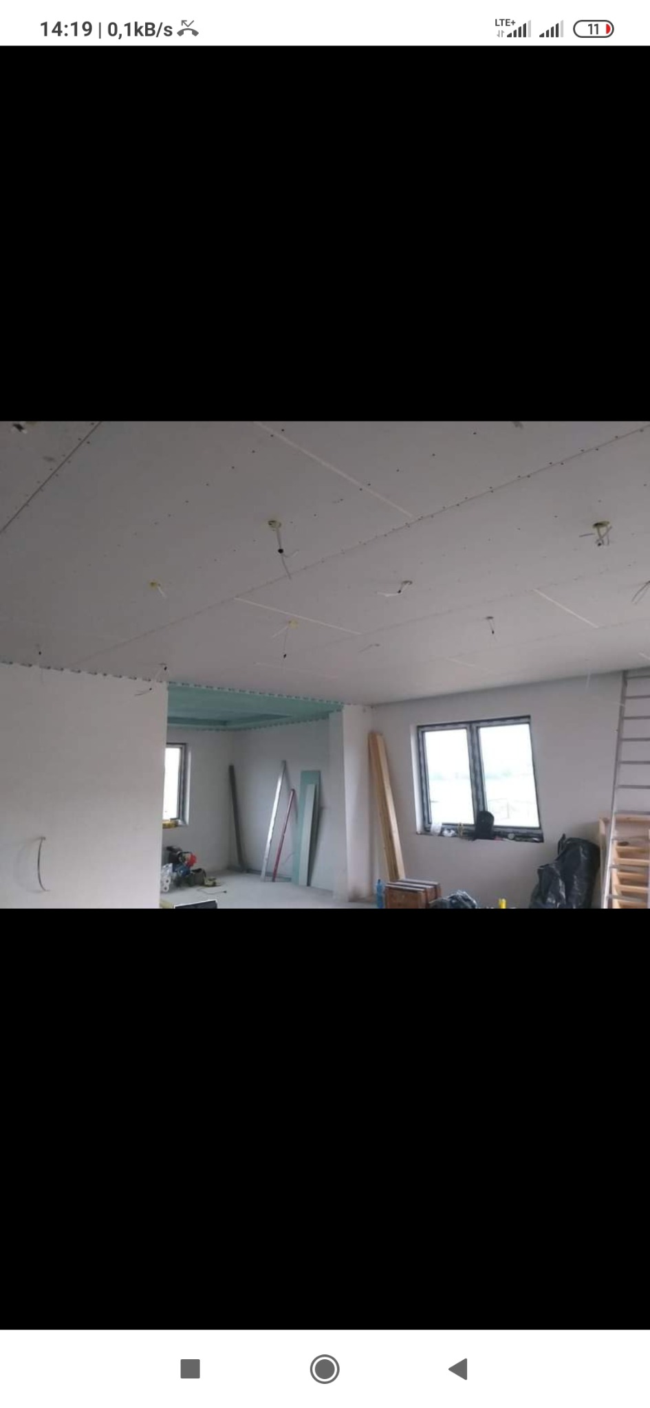 Widok wnętrza w trakcie prac wykończeniowych: zamontowany sufit podwieszany z płyt gipsowo-kartonowych z widocznymi przewodami oświetleniowymi, ściany przygotowane do malowania, okna z widokiem...