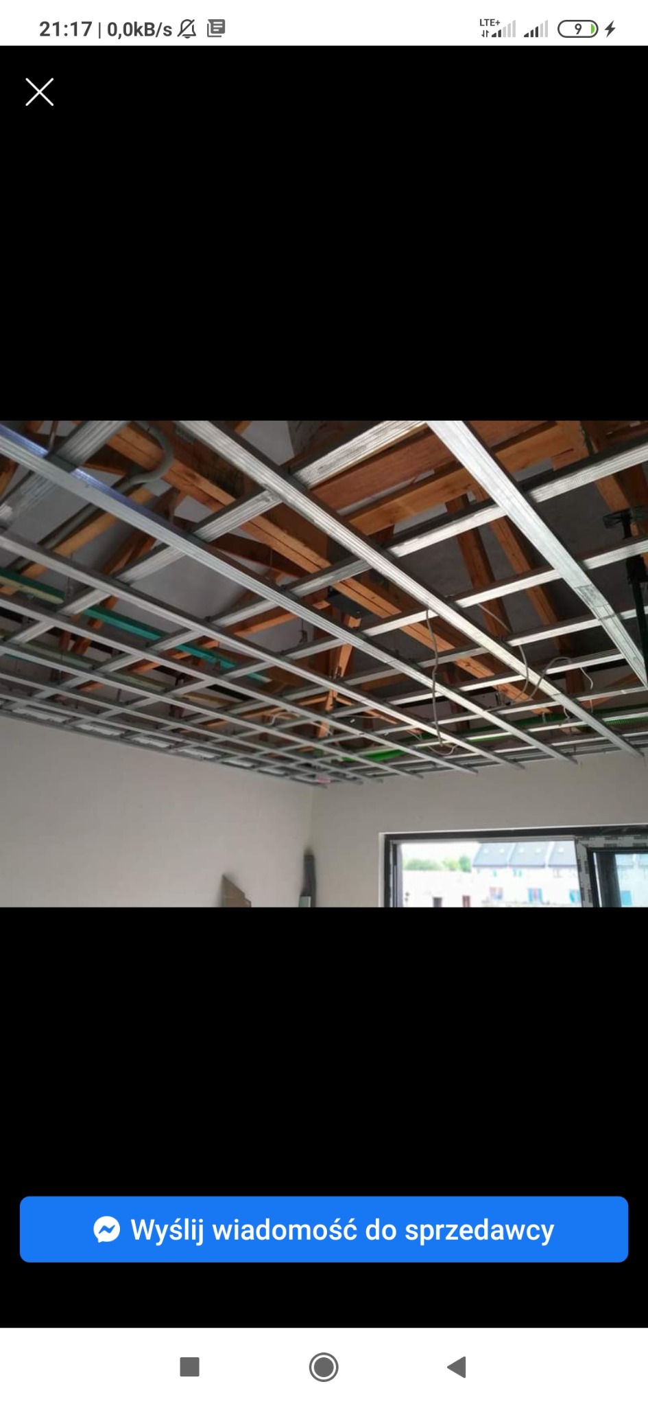 Konstrukcja metalowa podwieszanego sufitu zamontowana do drewnianych belek stropowych, widoczne instalacje, w tle jasna ściana i okno.