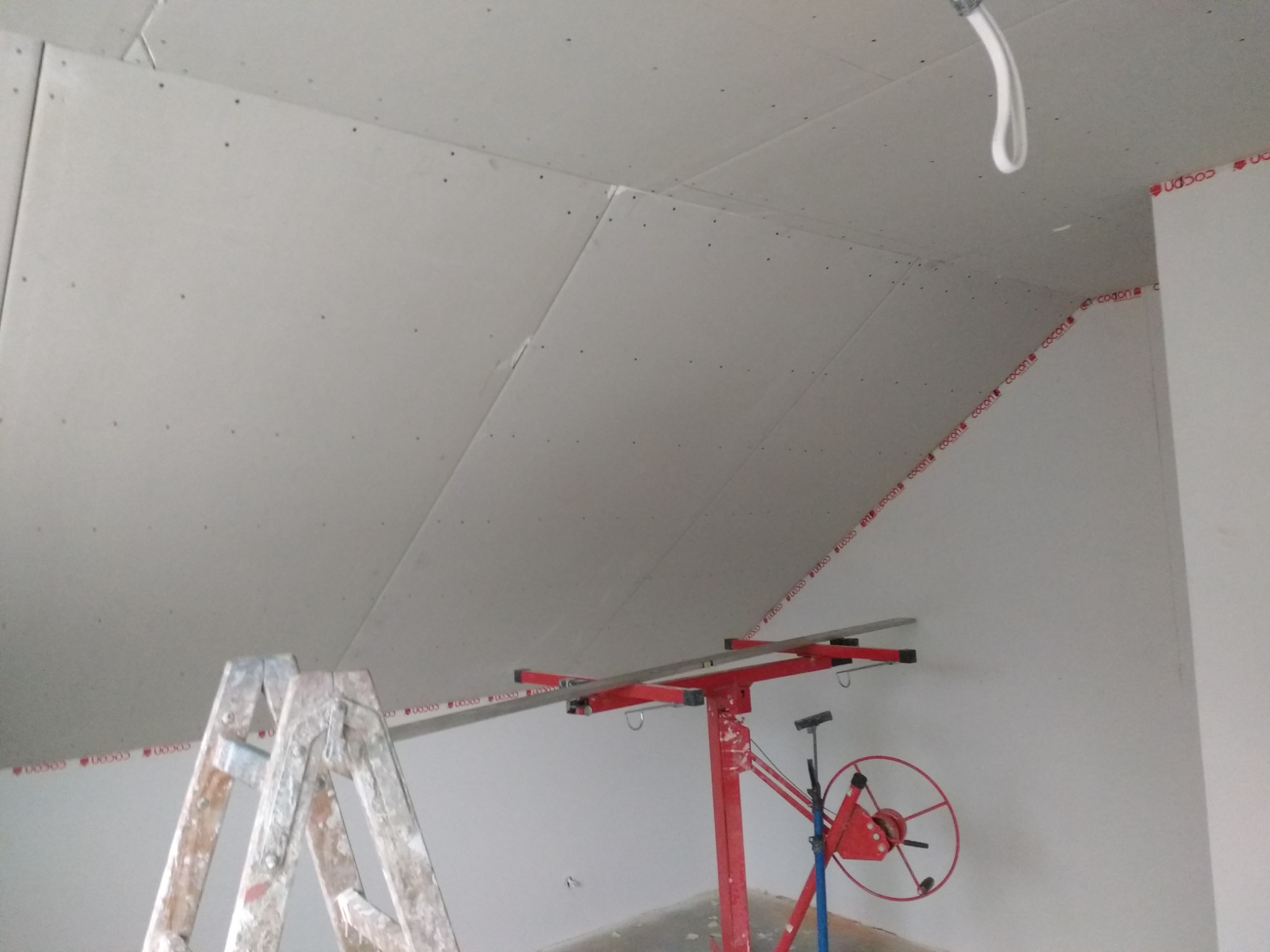 Montaż płyt na skośnym poddaszu z użyciem podnośnika, widoczna drabina i taśma miernicza z nadrukiem wzdłuż krawędzi ściany.