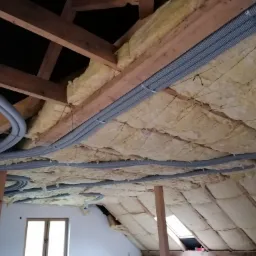 Ocieplone poddasze z widocznymi szarymi, karbowanymi rurami wentylacyjnymi przymocowanymi do drewnianej konstrukcji dachu.