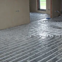 Instalacja ogrzewania podłogowego w trakcie montażu, widoczne rury grzewcze ułożone na siatce metalowej, pomieszczenie w stanie surowym, ściany bez wykończenia.