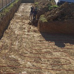 TOEKO Tobiasz Półchłopek - Instalacja rur ogrzewania podłogowego w wykopie ziemnym, widoczny budynek w tle i pracownicy przy wykopie, krajobraz pagórkowaty w oddali.