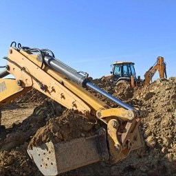 TOEKO Tobiasz Półchłopek - Żółta koparka Cukurova zagłębia się łyżką w ziemię, w tle inne maszyny budowlane i błękitne niebo. Prace ziemne w toku.