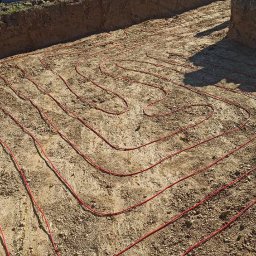 TOEKO Tobiasz Półchłopek - Czerwone rury ogrzewania podłogowego ułożone w wykopie, widok z góry. W tle zielone pola i ogrodzenie.