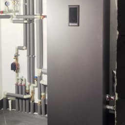 TOEKO Tobiasz Półchłopek - Szara pompa ciepła z ekranem dotykowym, otoczona rurami z izolacją, zamontowana w pomieszczeniu z szarą podłogą. Widoczny czerwony zbiornik wyrównawczy.