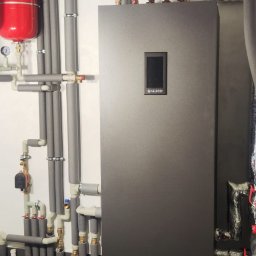 TOEKO Tobiasz Półchłopek - Nowoczesna instalacja grzewcza z szarą pompą ciepła, czerwonym zbiornikiem wyrównawczym i izolowanymi rurami na tle białej ściany i szarej podłogi.