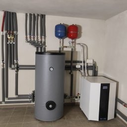 TOEKO Tobiasz Półchłopek - Szary zbiornik buforowy z manometrem, biała pompa ciepła i instalacja rurowa z izolacją w pomieszczeniu technicznym na tle beżowych płytek podłogowych.