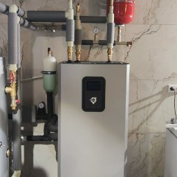 TOEKO Tobiasz Półchłopek - Szara pompa ciepła Thermia z podłączonymi rurami i zbiornikiem wyrównawczym na tle marmurowej ściany i podłogi. Widoczne elementy instalacji wentylacyjnej pod sufitem.