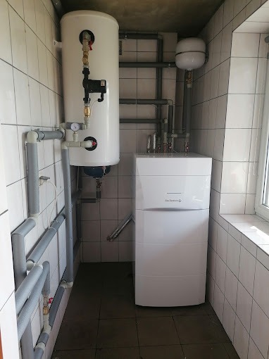 Biały, kondensacyjny piec gazowy Electrolux z zasobnikiem wody w kotłowni wyłożonej białymi płytkami. Widoczne rury i zawory instalacji grzewczej.