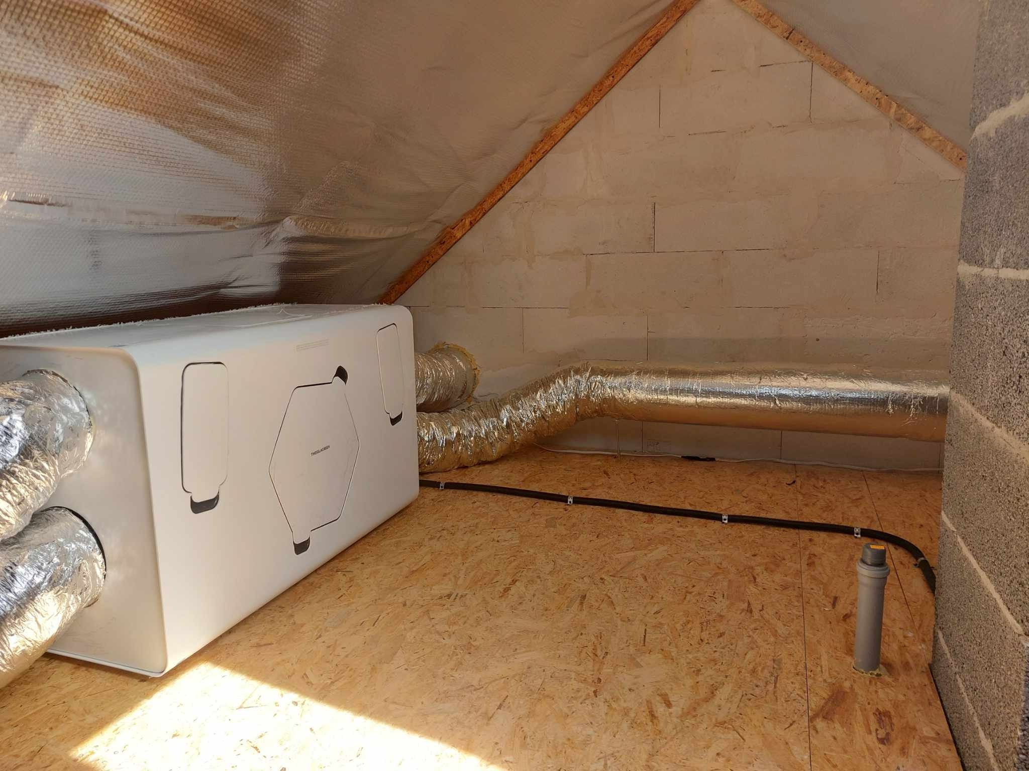 Biała jednostka rekuperacji z podłączonymi przewodami wentylacyjnymi w przestrzeni poddasza z drewnianą podłogą i izolacją. Widok na instalację w surowym stanie.