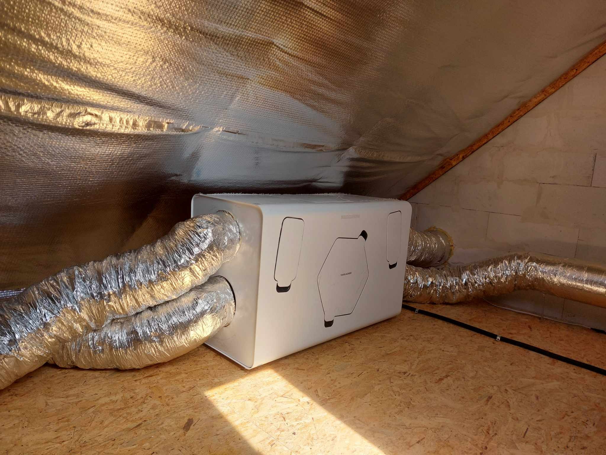 Centrala rekuperacyjna z podłączonymi przewodami wentylacyjnymi w pomieszczeniu ze skośnym sufitem wykończonym folią aluminiową. Widoczna podłoga z płyty OSB.