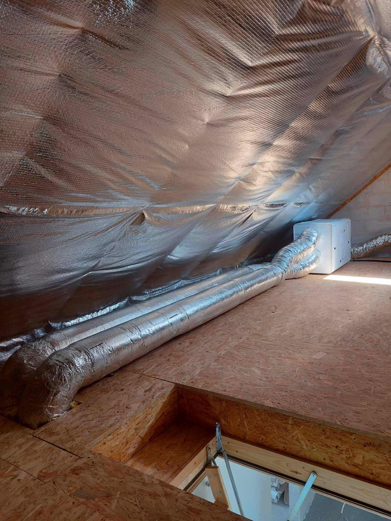 Dwie srebrne rury wentylacyjne prowadzą do białej centrali rekuperacyjnej na poddaszu z drewnianą podłogą i izolacją termiczną na skośnym dachu.