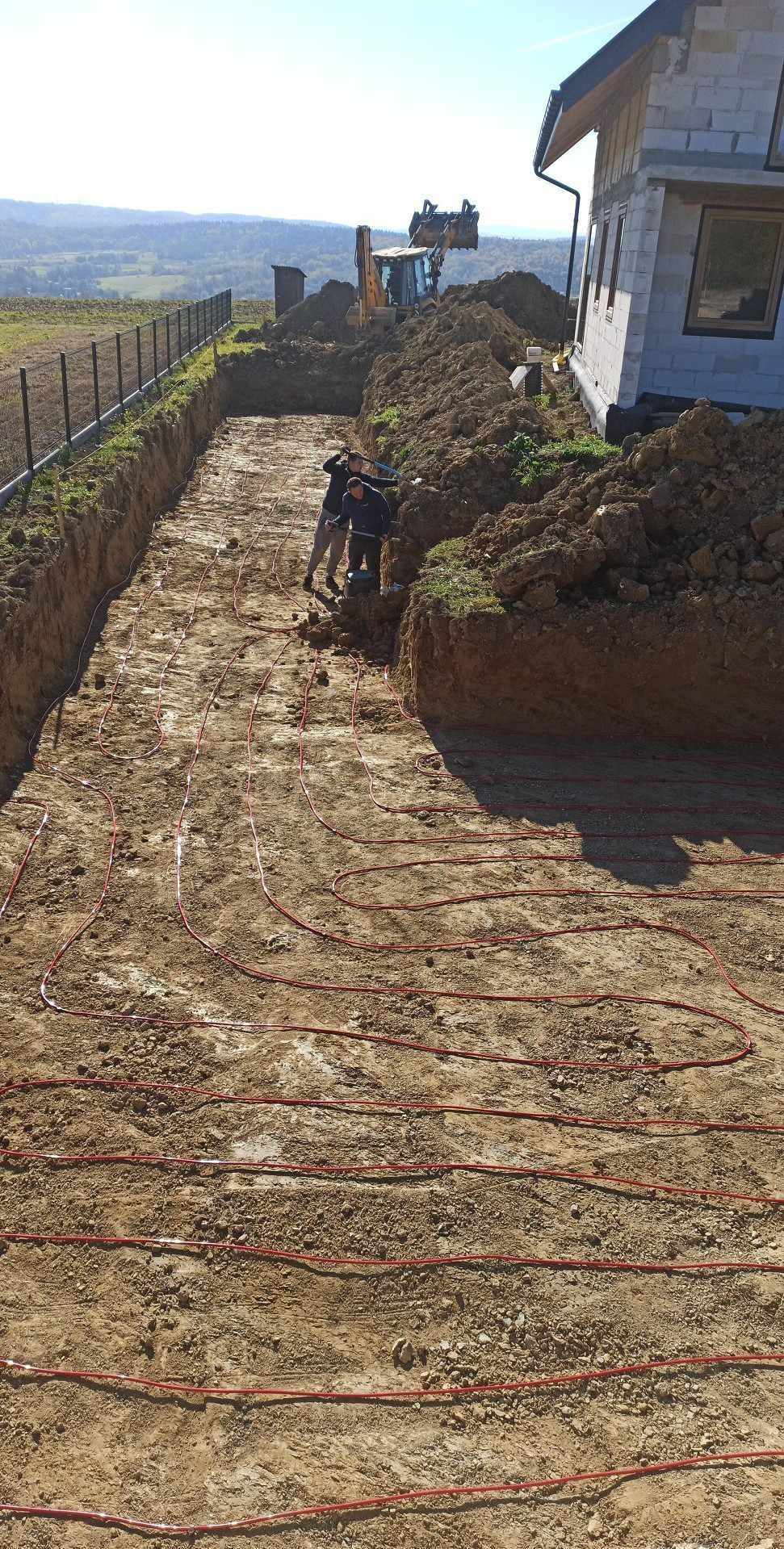 Instalacja rur ogrzewania podłogowego w wykopie ziemnym, widoczny budynek w tle i pracownicy przy wykopie, krajobraz pagórkowaty w oddali.