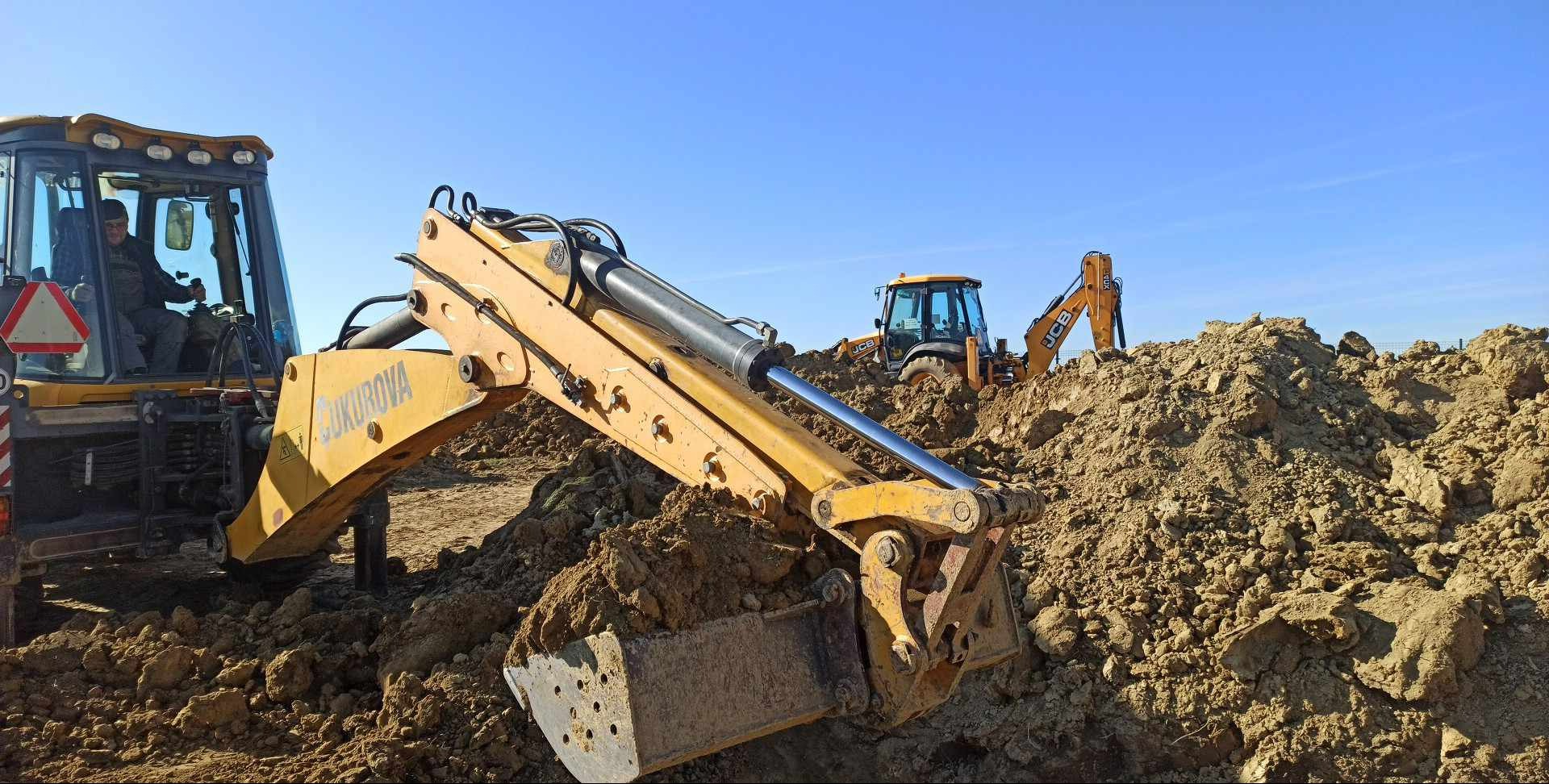 Żółta koparka Cukurova zagłębia się łyżką w ziemię, w tle inne maszyny budowlane i błękitne niebo. Prace ziemne w toku.