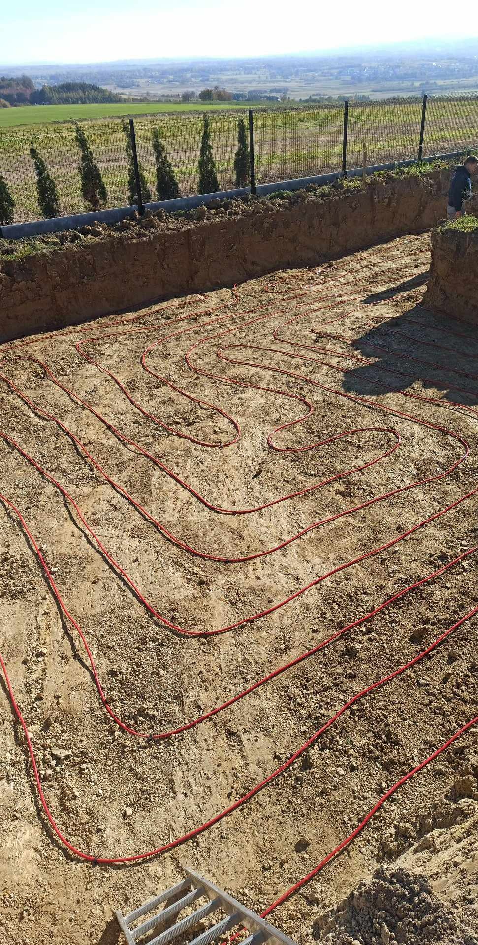 Czerwone rury ogrzewania podłogowego ułożone w wykopie, widok z góry. W tle zielone pola i ogrodzenie.