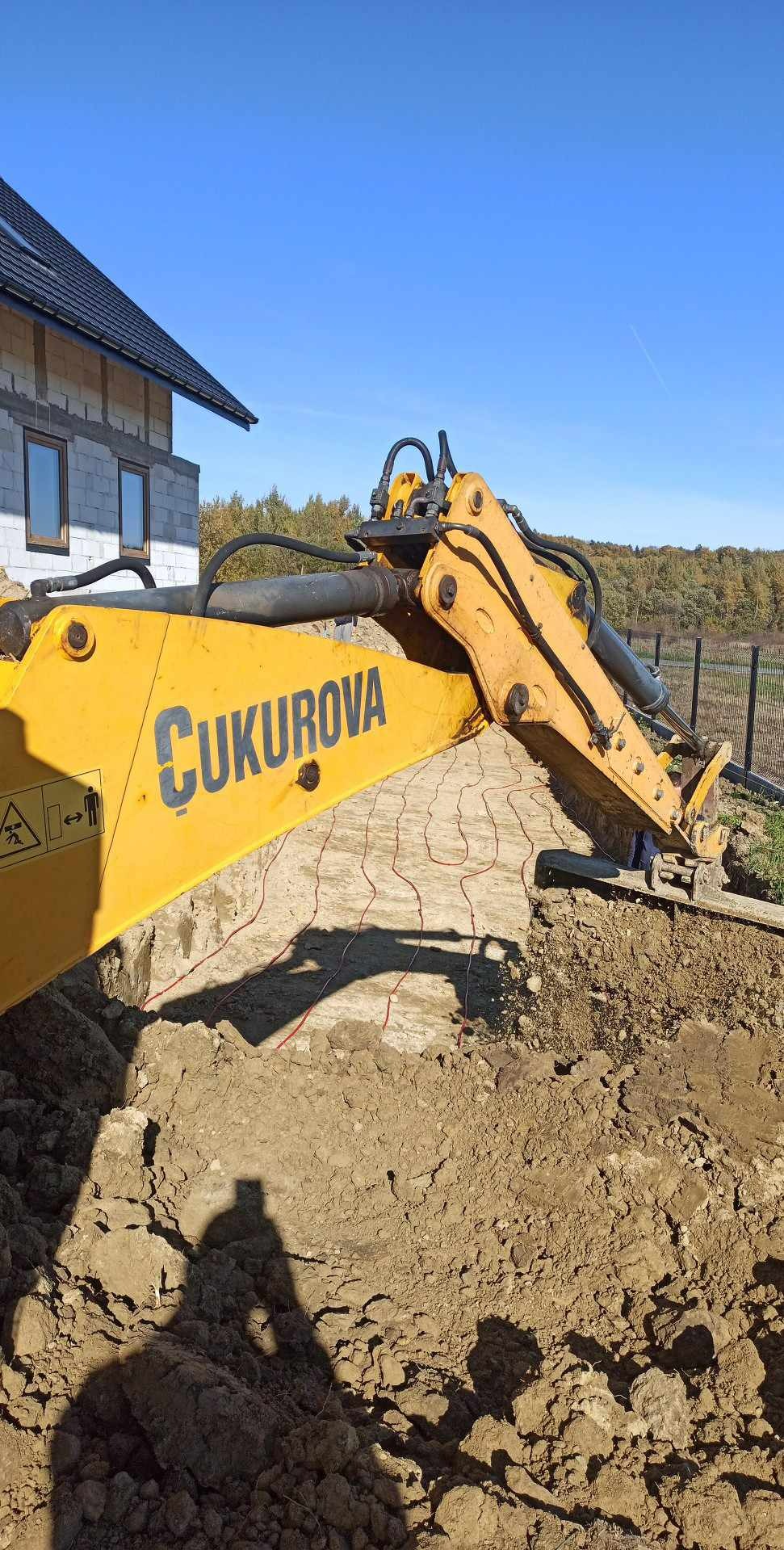 Żółta koparka Cukurova przygotowuje teren pod budowę domu jednorodzinnego. Widoczne przewody w wykopie, ogrodzenie i drzewa w tle.