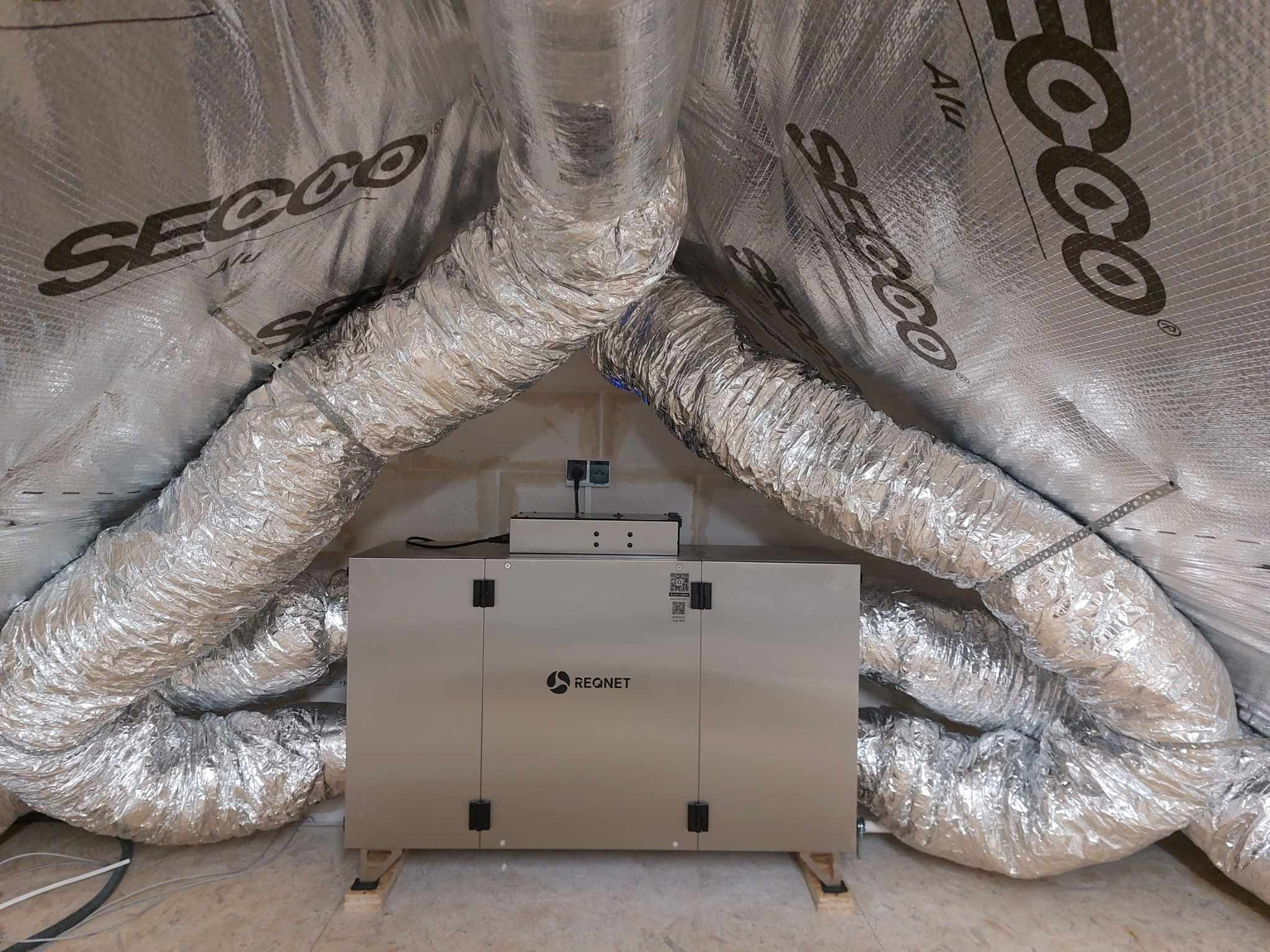 Szara centrala wentylacyjna REQNET z podłączonymi, elastycznymi przewodami wentylacyjnymi w izolowanym pomieszczeniu na poddaszu. Widoczne elementy montażowe.