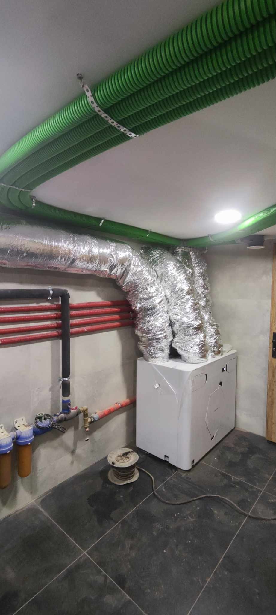 Zakończona instalacja rekuperacji z zielonymi rurami wentylacyjnymi pod sufitem i srebrnymi, izolowanymi przewodami do białej centrali wentylacyjnej.
