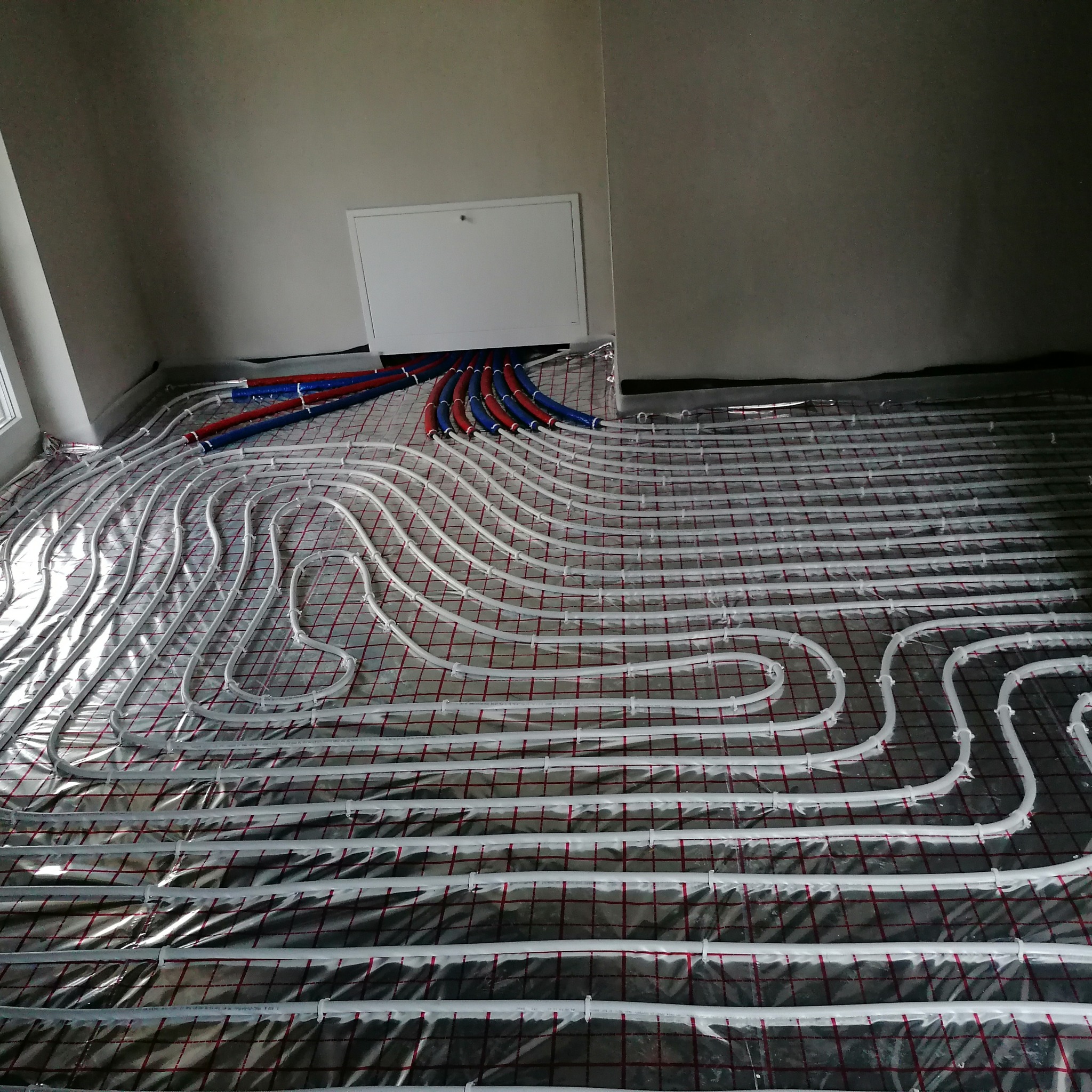 Ułożone białe rury ogrzewania podłogowego na folii termoizolacyjnej z czerwoną siatką, widoczne niebieskie i czerwone rury doprowadzające, biała klapa rewizyjna.