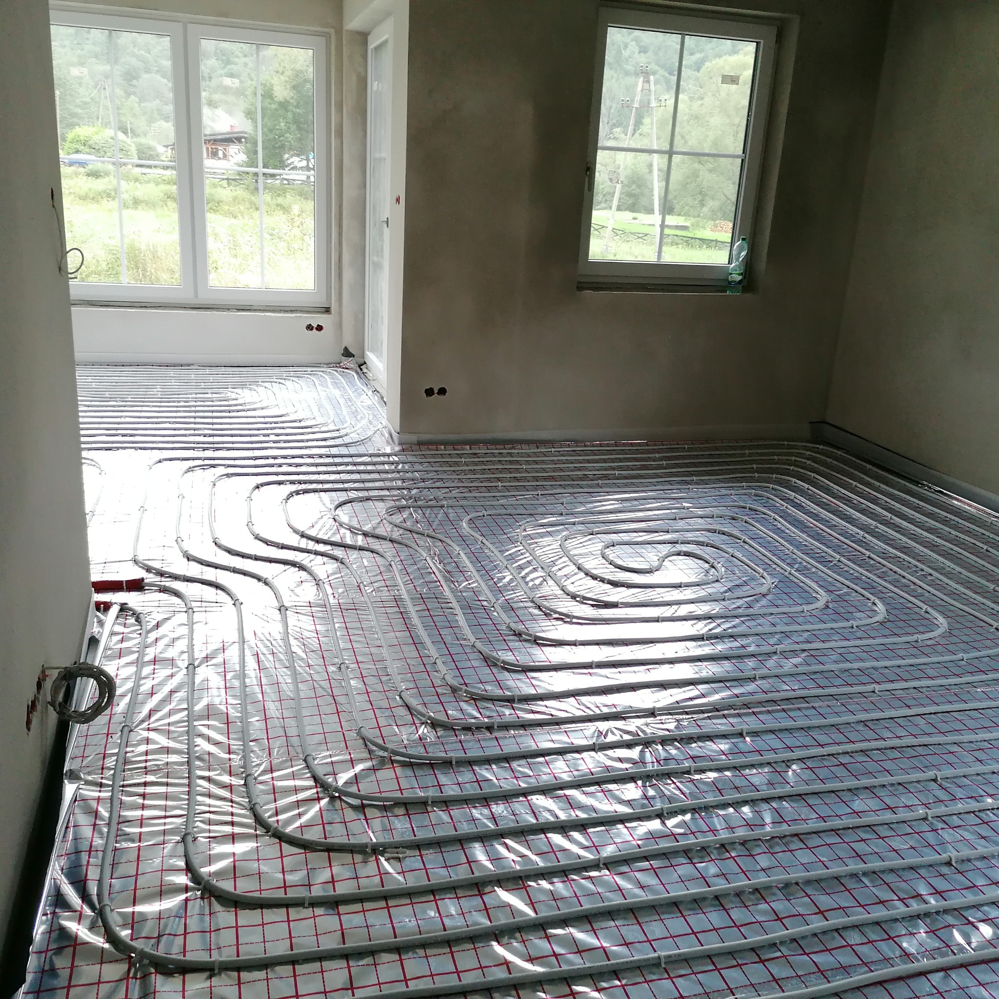 Ułożone rury ogrzewania podłogowego na folii izolacyjnej w jasnym, surowym wnętrzu z oknami wychodzącymi na zieleń. Widoczne detale instalacji i przygotowanie do wylewki.