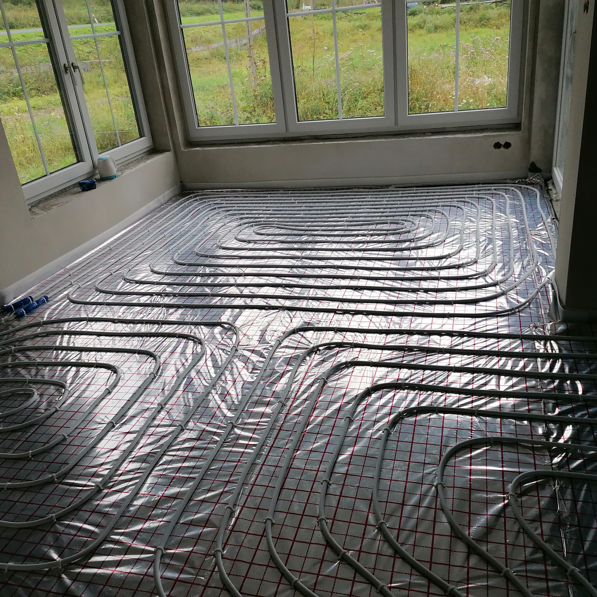 Szare rury ogrzewania podłogowego ułożone na srebrnej folii z czerwoną siatką w jasnym pomieszczeniu z dużymi oknami. Widoczne złącza rur.