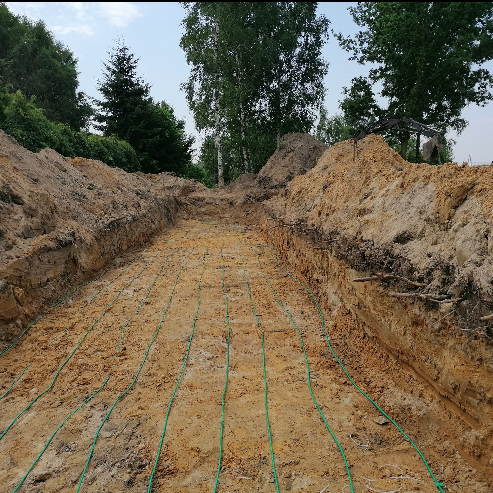 Wykop pod instalację z zielonymi rurami, prawdopodobnie gruntowy wymiennik ciepła. Widoczne ściany wykopu z ziemi i drzewa w tle.