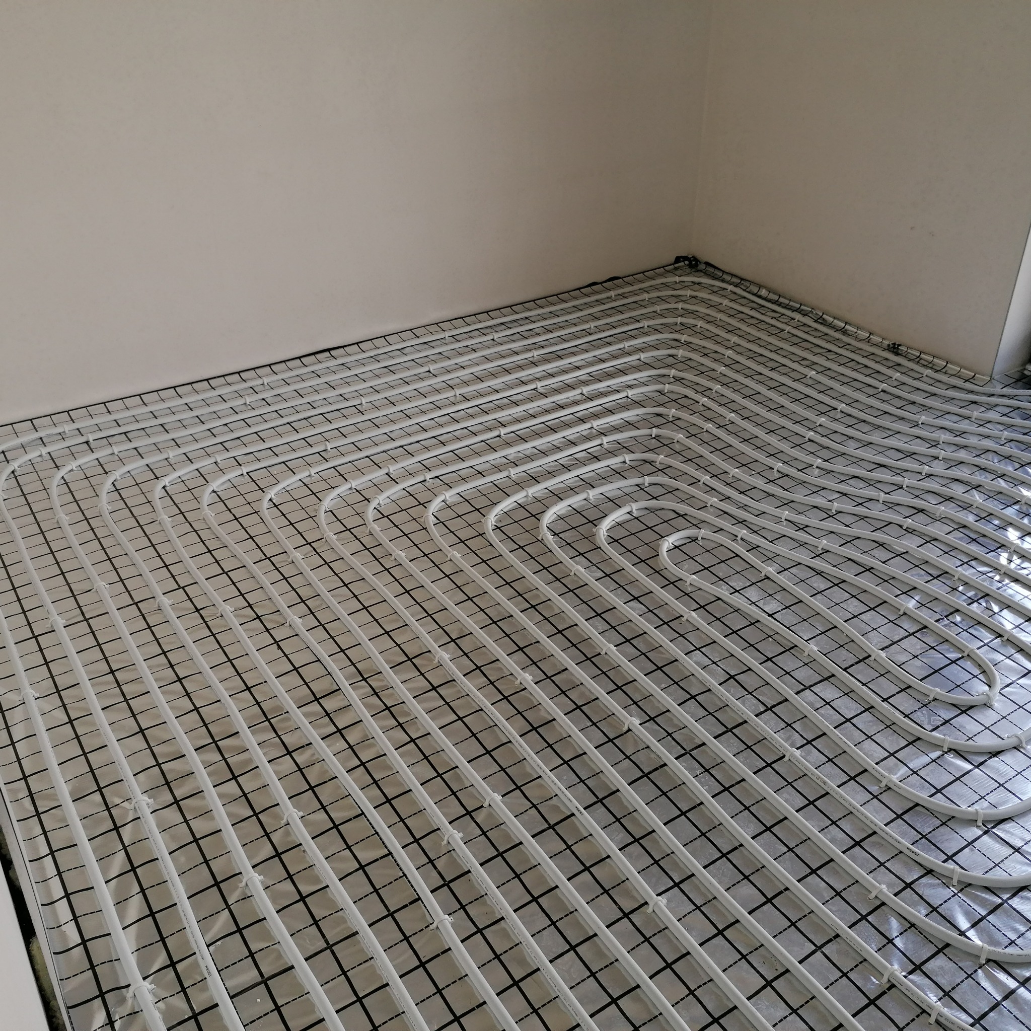 Białe rury ogrzewania podłogowego ułożone na folii z czarną siatką w rogu jasnego pomieszczenia, widok z góry, etap instalacji przed wylewką.