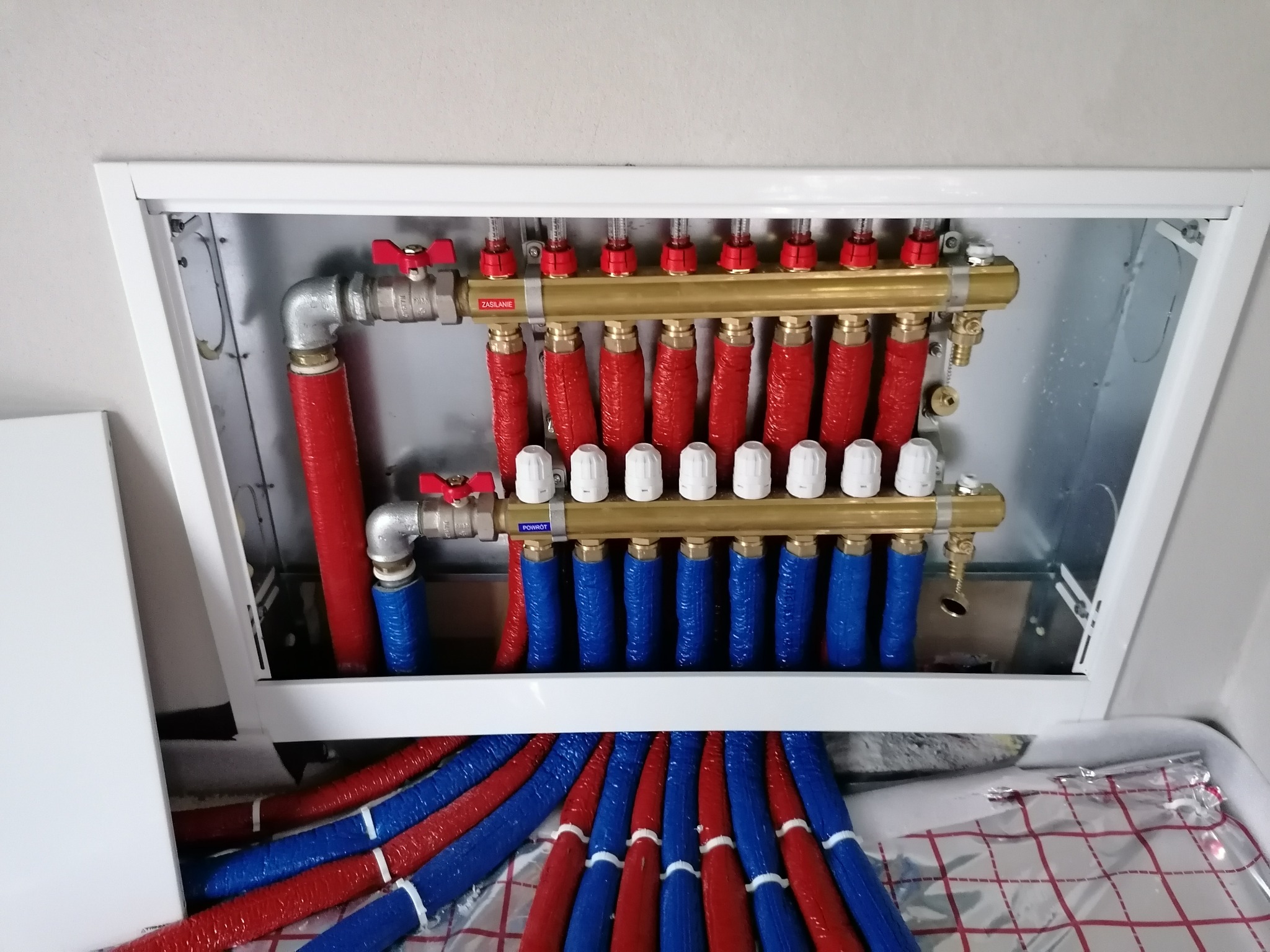 Rozdzielacz ogrzewania podłogowego w białej obudowie, z czerwonymi i niebieskimi rurami, widoczne zawory i elementy instalacji.