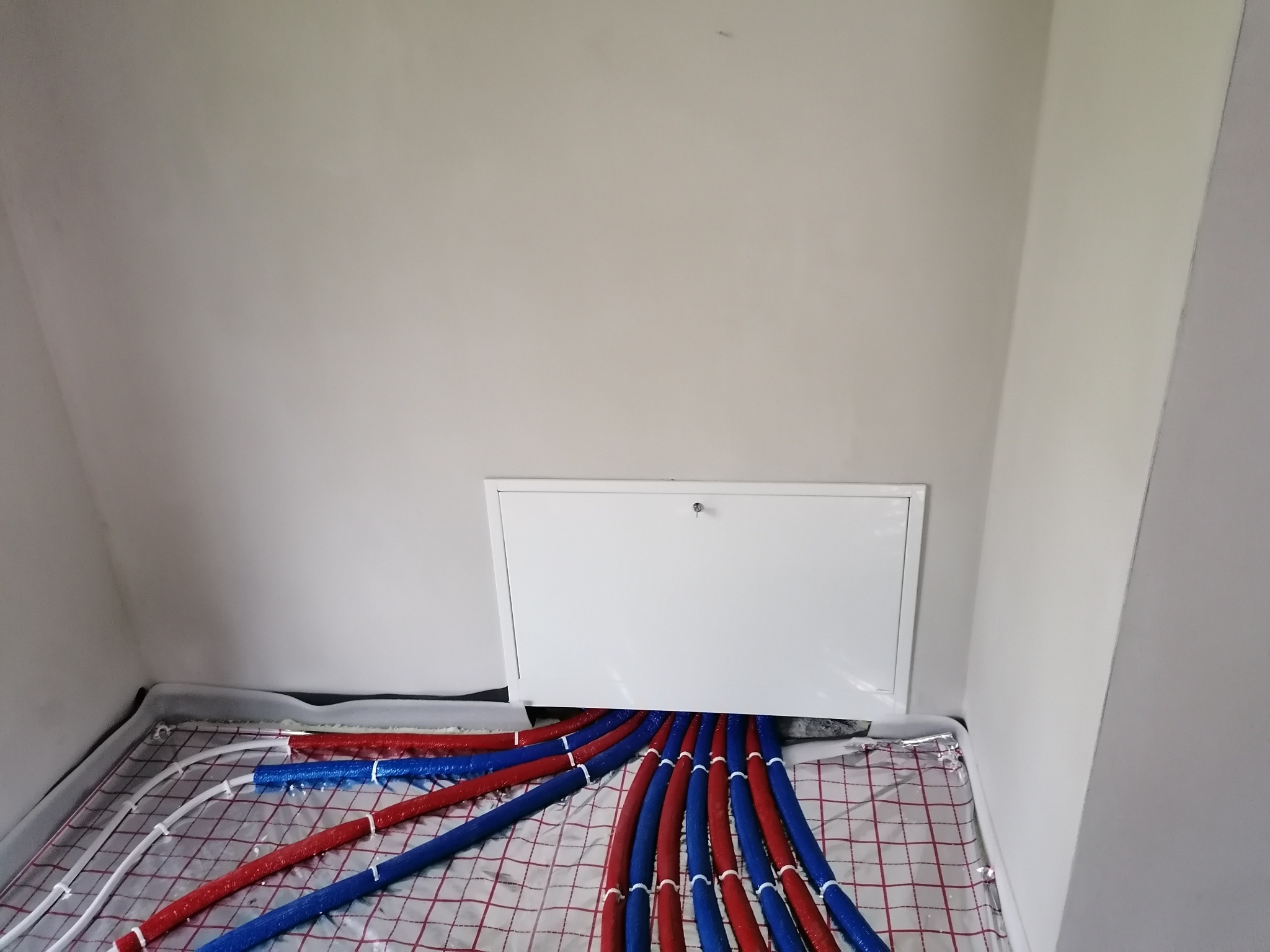 Rozłożone rury ogrzewania podłogowego w izolacji czerwono-niebieskiej, wychodzące spod białej klapy rewizyjnej na tle białych ścian poddasza.