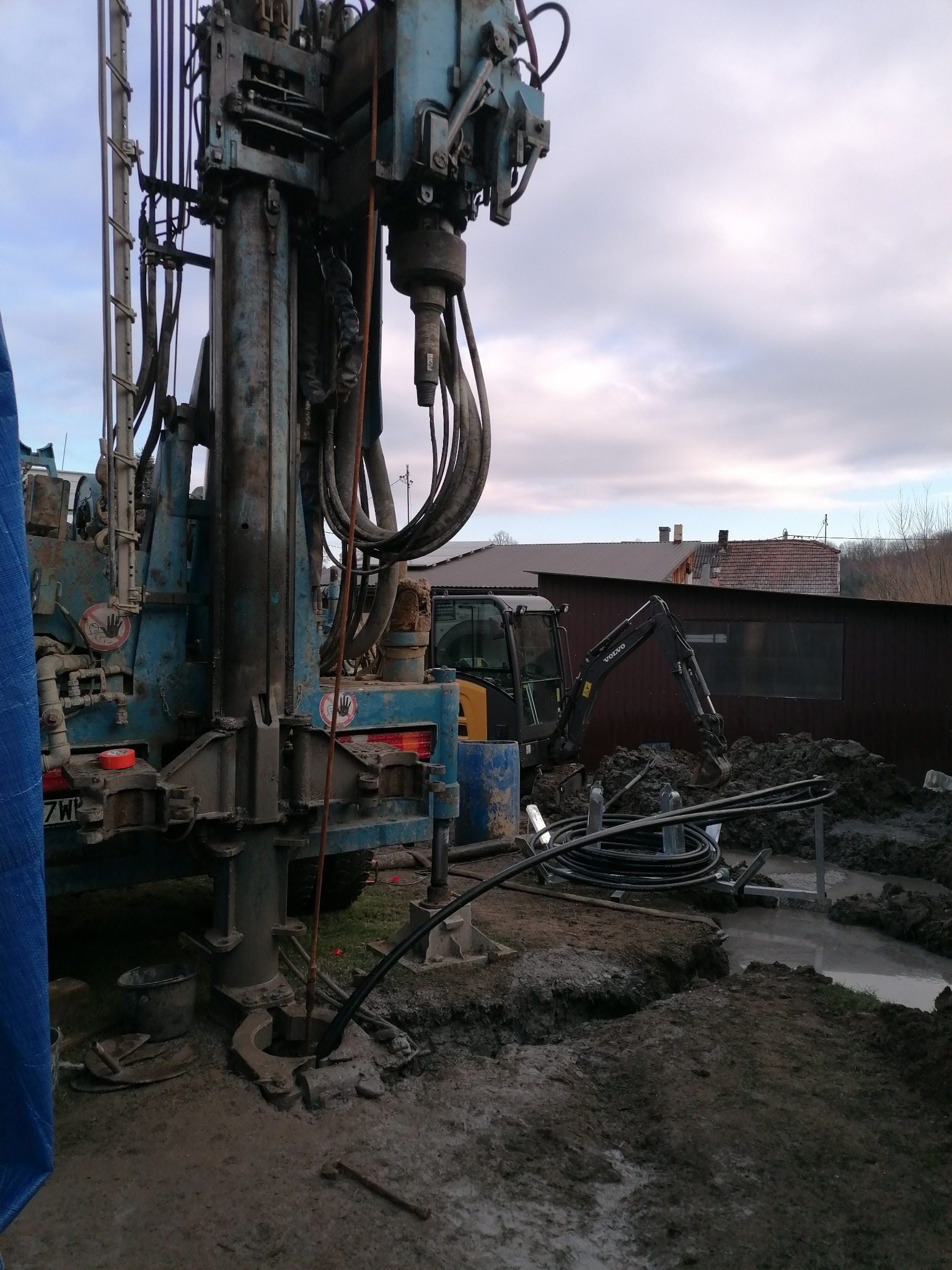 Wiertnica w trakcie pracy na zewnątrz, z widocznym małym koparką w tle i błotnistym terenem. Urządzenie wiertnicze w kolorze niebieskim, z licznymi przewodami i elementami konstrukcyjnymi.