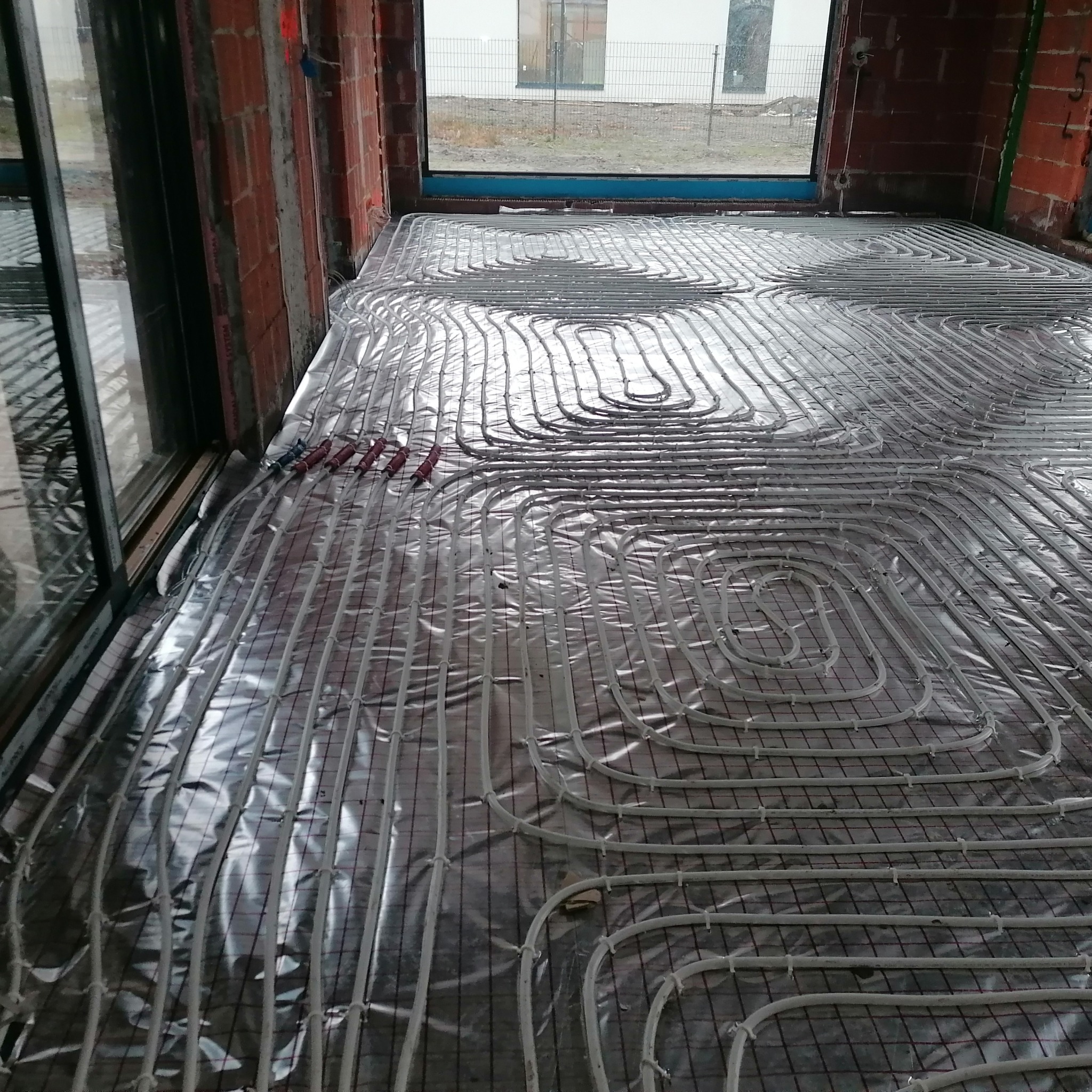 Ułożone rury ogrzewania podłogowego na folii izolacyjnej, widoczne okno i surowe ściany z cegły. Instalacja w trakcie realizacji, przed wylaniem posadzki.