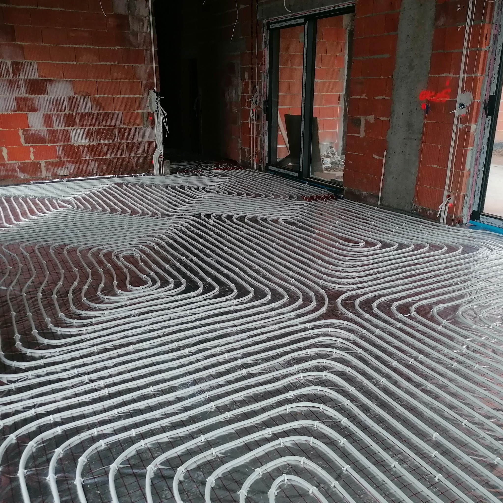 Białe rury ogrzewania podłogowego ułożone spiralnie na folii w Krościenko Wyżne, widok z góry. Instalacja w surowym wnętrzu z ceglanymi ścianami.