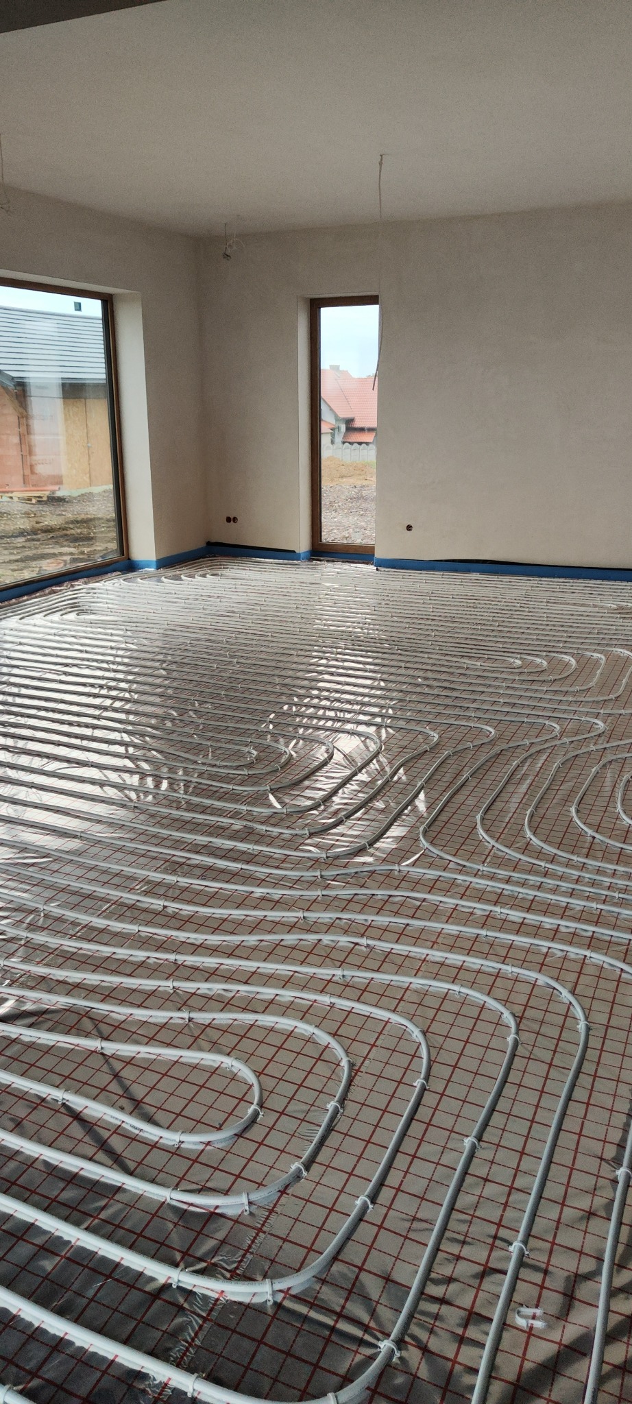 Białe rury ogrzewania podłogowego ułożone na folii z czerwoną kratką w jasnym pomieszczeniu z oknami i drzwiami balkonowymi. Widoczne nieotynkowane ściany.