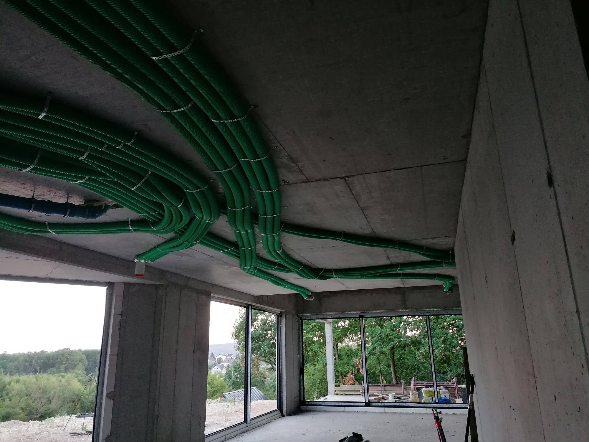 Zielone rury wentylacyjne przymocowane do surowego betonowego sufitu w budynku w stanie deweloperskim z dużymi oknami i widokiem na zieleń.